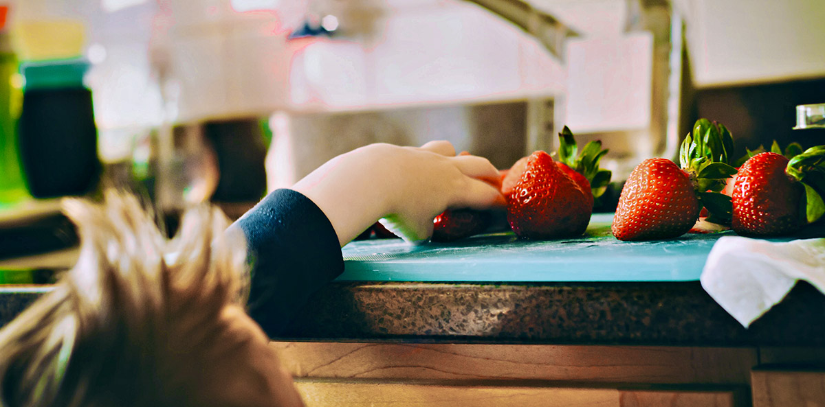 Young boy stretches a head up onto kitchen counter to grab a luscious red strawberry.