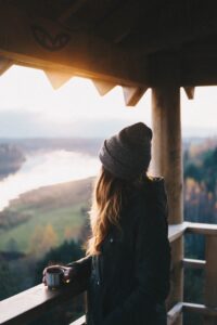 Photo of woman in wool cap and heavy coat drinking coffee on a timbered porch and looking out at a mountain landscape.